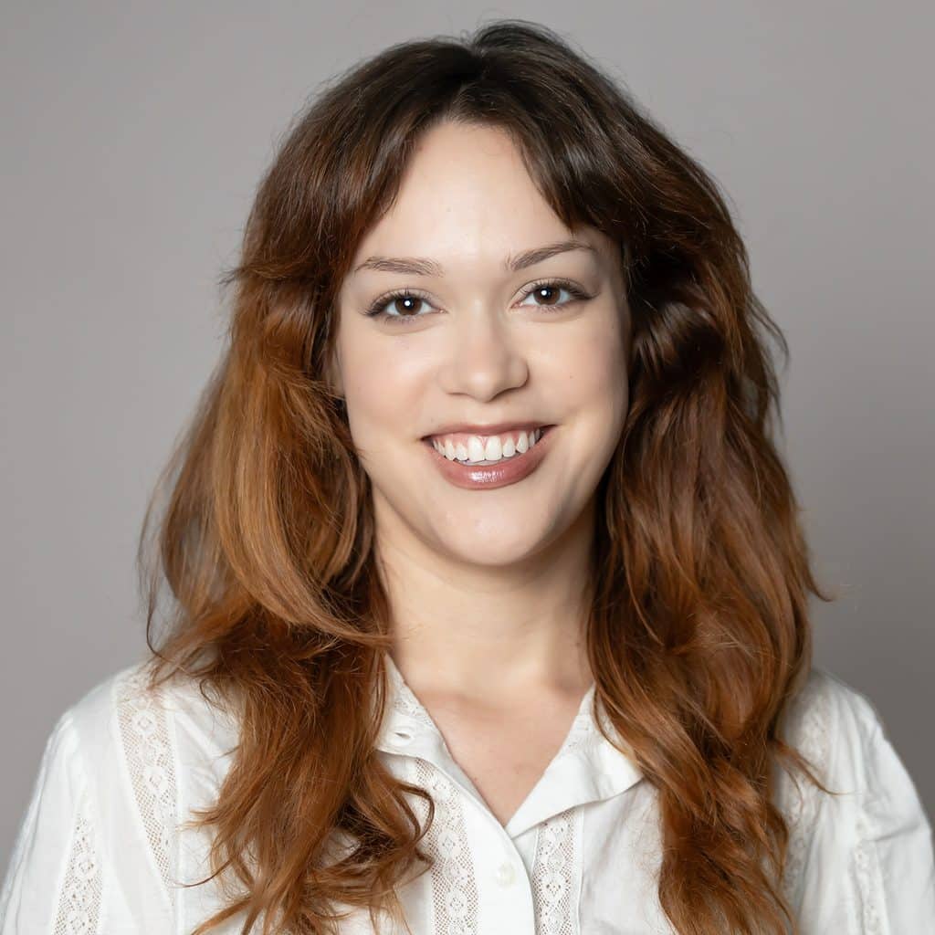A headshot of a smiling woman with long brown hair wearing a white collared shirt.