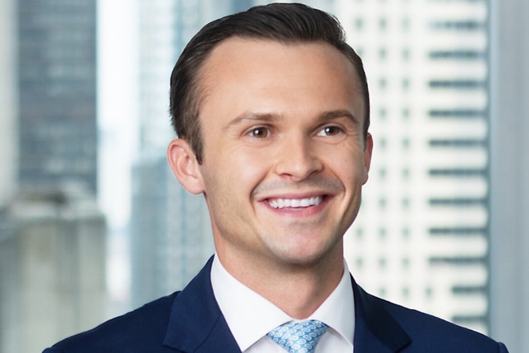 A man in a dark blue suit and light blue tie is smiling and looking slightly to the side. He is standing indoors in front of large windows with tall office buildings visible outside.