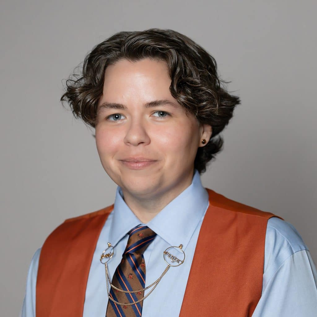 A person with short, wavy brown hair wears a light blue dress shirt, a striped necktie with a gold collar chain, and an orange vest.