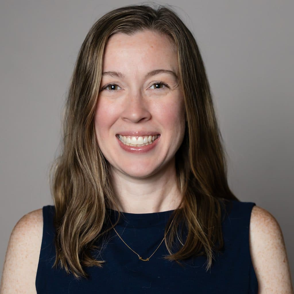 A woman with shoulder-length brown hair wearing a dark blue sleeveless top and a gold necklace smiles at the camera.