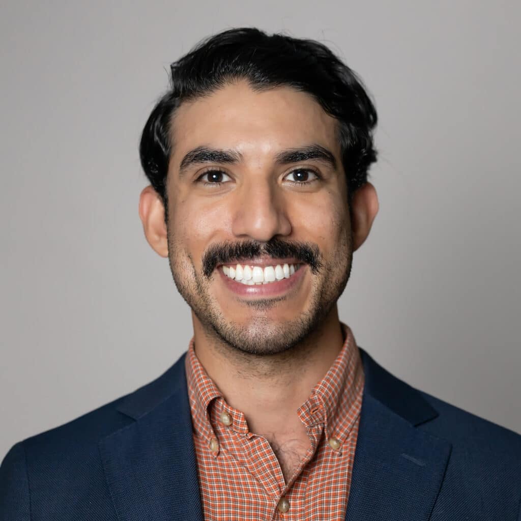 A man with a mustache smiling in a navy blazer and orange checkered shirt.