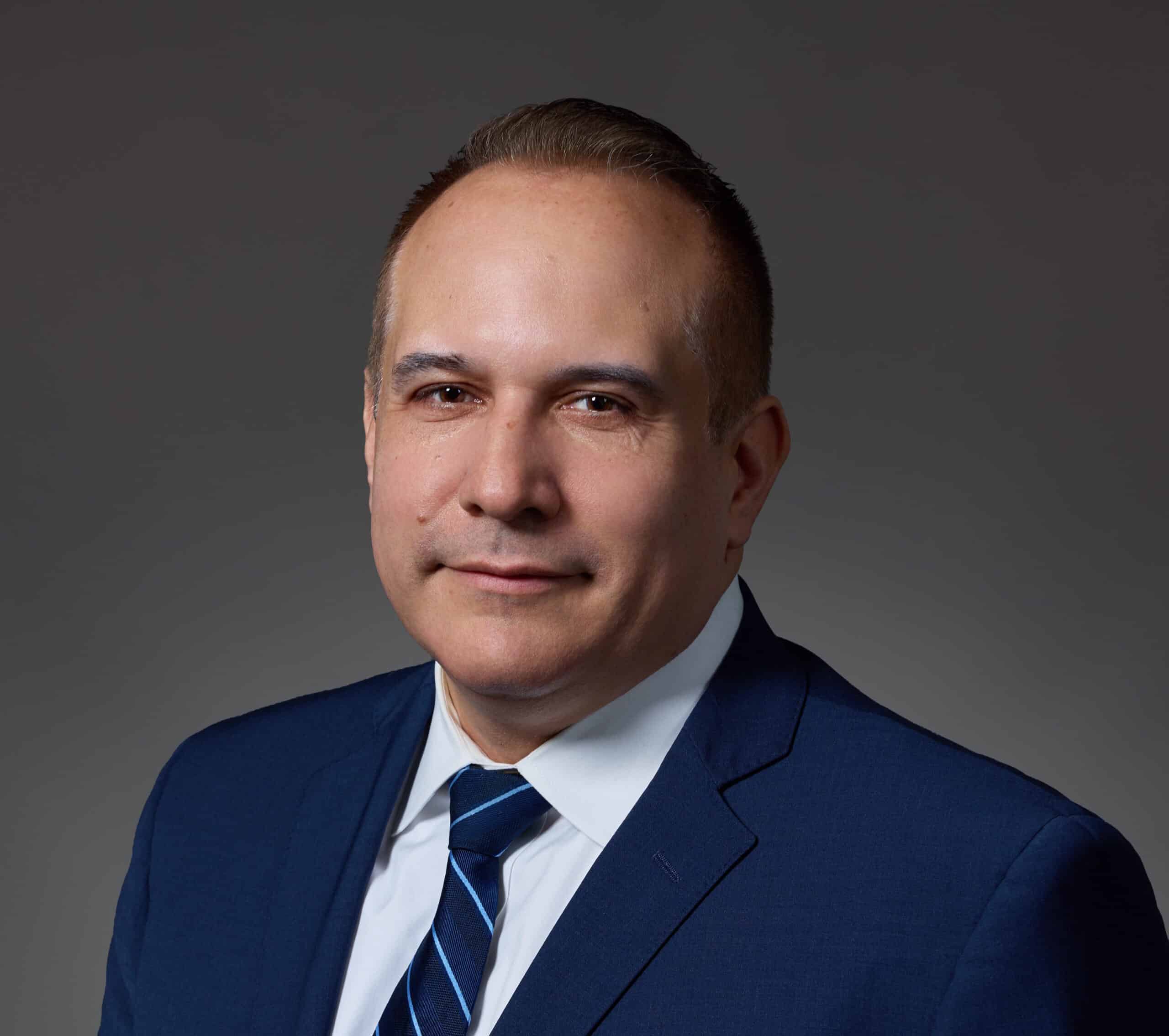A man in a blue suit, white shirt, and striped tie stands against a plain gray background, facing forward with a neutral expression.