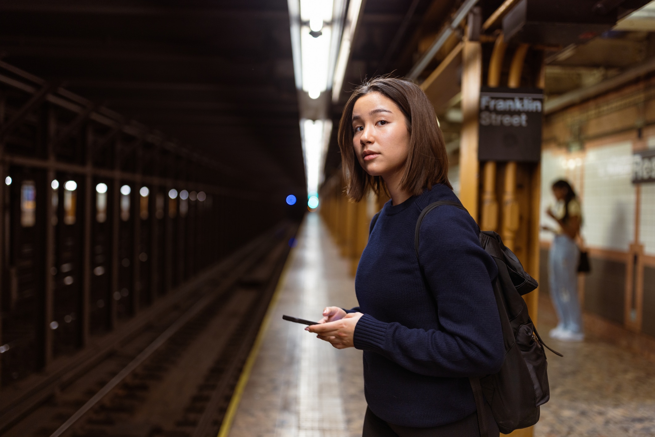 A woman with a backpack stands on a subway platform holding a phone. She looks toward the camera. Behind her, another person stands near a wall with a Franklin Street sign. The train tracks are visible.