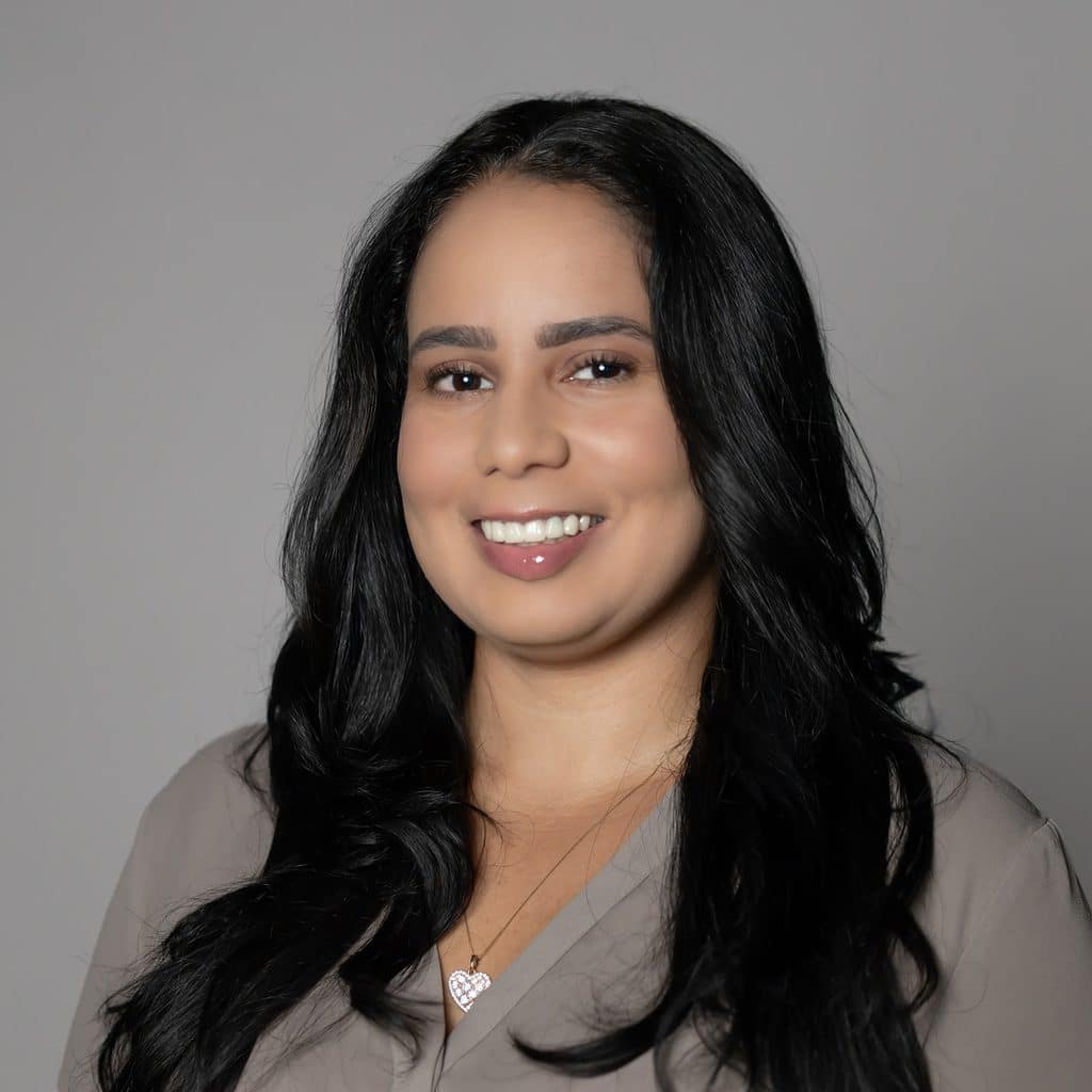 A professional headshot of a woman with long, dark hair smiling against a grey background.