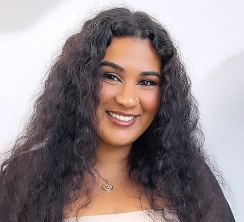 A woman with long, dark, curly hair smiles at the camera. She is wearing a light-colored top, a black cardigan, and a gold necklace with a pendant. The background is plain and light-colored.