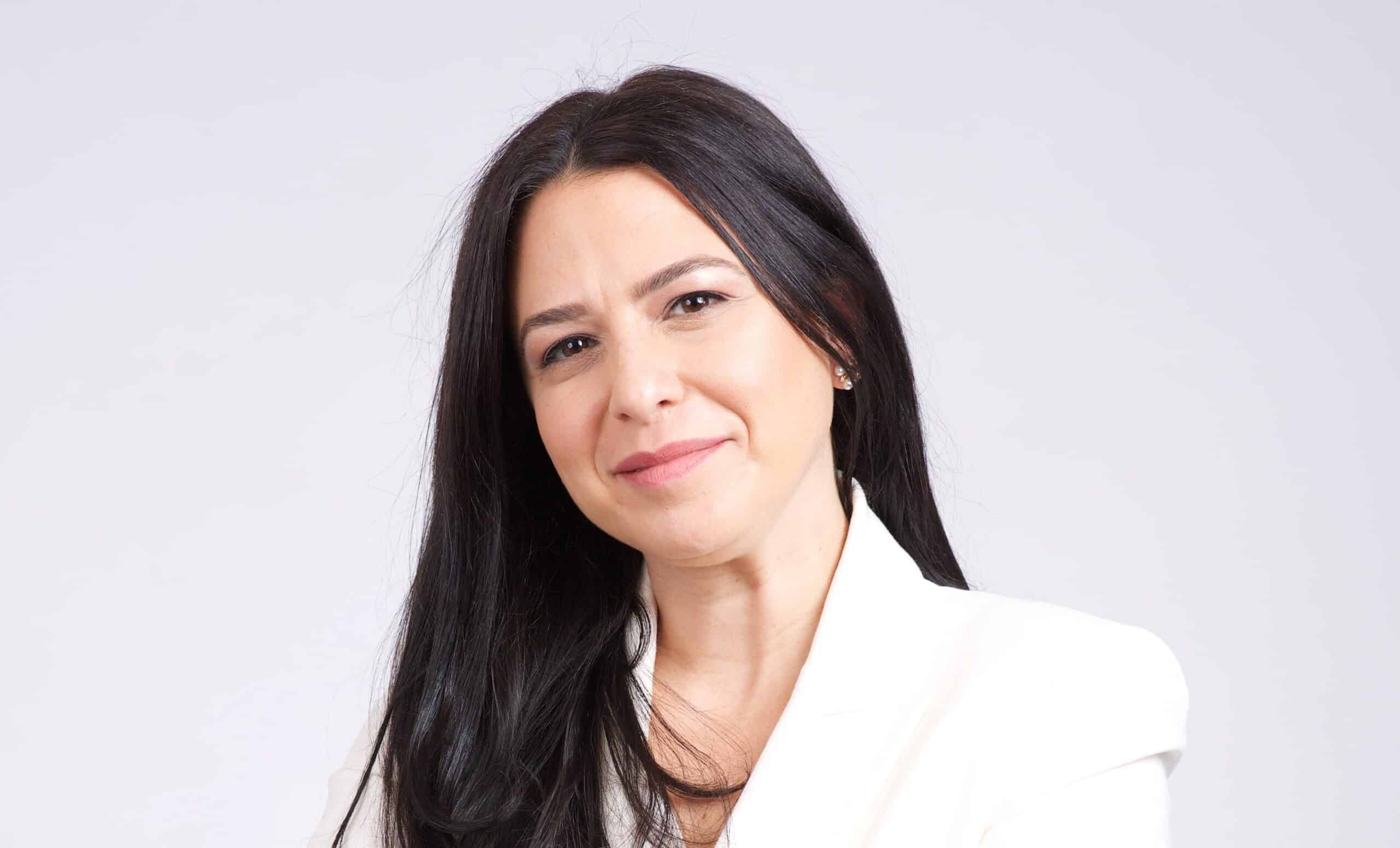 A woman with long black hair, wearing a white blazer and black pants, stands against a plain white background with her arms crossed, looking at the camera with a slight smile.