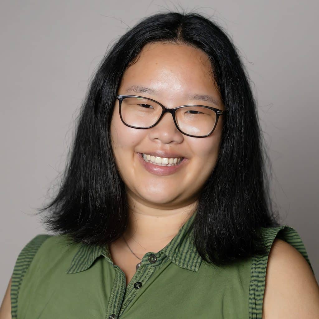 A young person with shoulder-length dark hair and glasses smiles at the camera, wearing a green collared shirt.