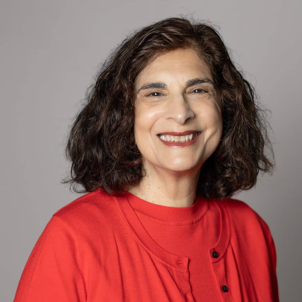 A smiling woman with shoulder-length dark curly hair wearing a red shirt.