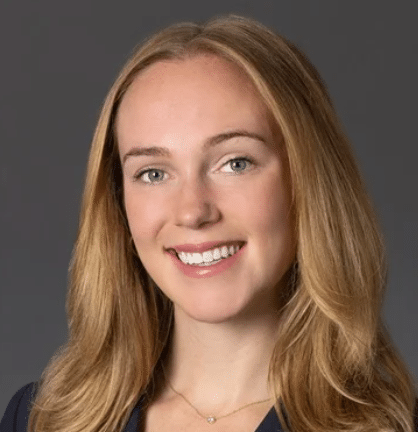 A woman with long, light brown hair wears a dark blazer over a white top and a delicate necklace. She is smiling at the camera against a plain gray background.
