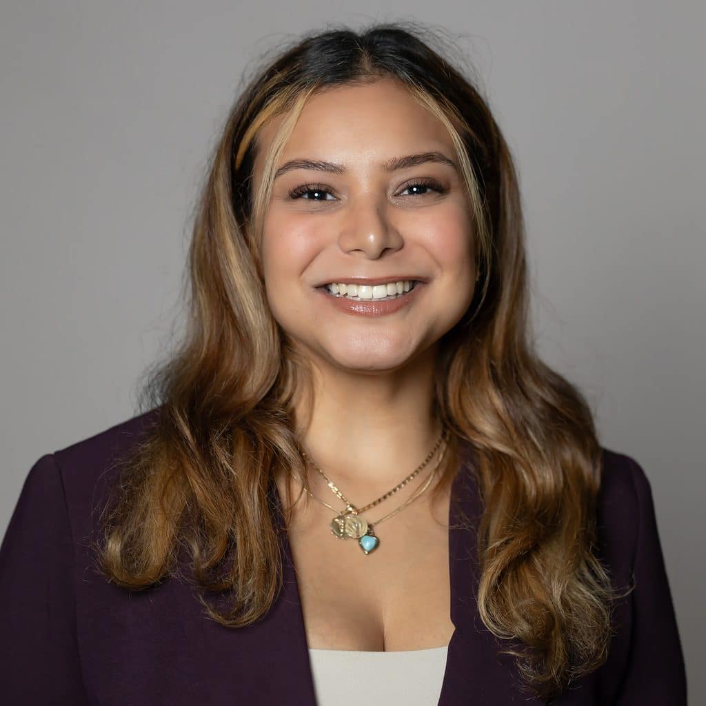 A smiling woman with shoulder-length highlighted hair wearing a dark purple blazer and layered necklaces.