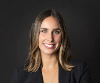 A woman with shoulder-length brown hair, wearing a black blazer and a necklace, smiles at the camera against a plain dark background.