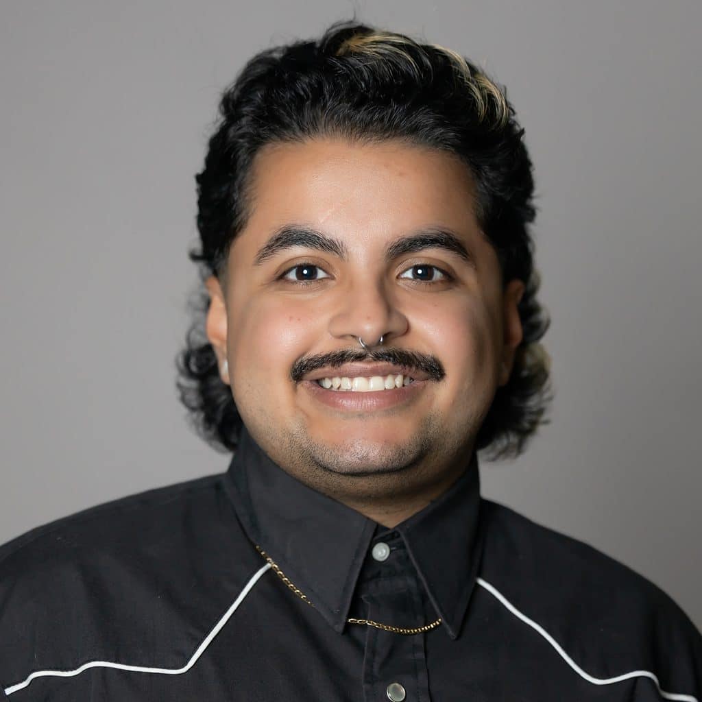 A portrait of a person with a mustache and septum piercing, smiling at the camera.