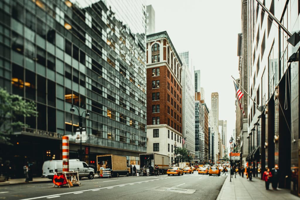 City street view with tall buildings, yellow taxis, trucks, and cars. The sidewalks have a few people, and American flags are visible on the right. The atmosphere appears busy with urban activity.