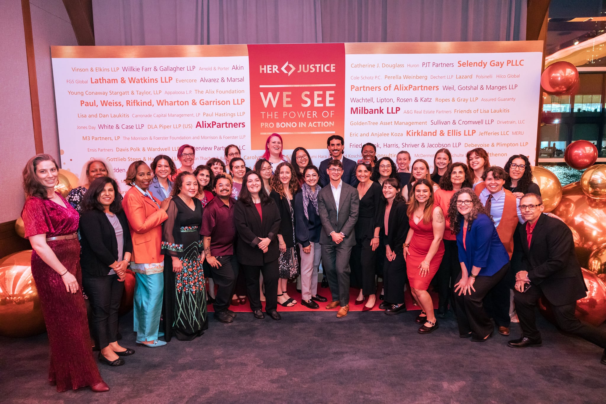 A group of people stand and pose for a photo at a Her Justice event, with sponsor boards and large red and gold ornaments in the background. Most attendees are dressed in business or semi-formal attire.
