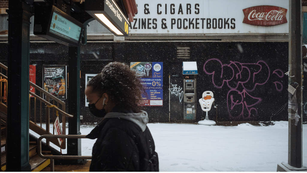 A person wearing a mask walks by a snow-covered street, with graffiti, posters, and a snowman figure in front of a closed shop with a Coca-Cola sign above. Snow is falling.
