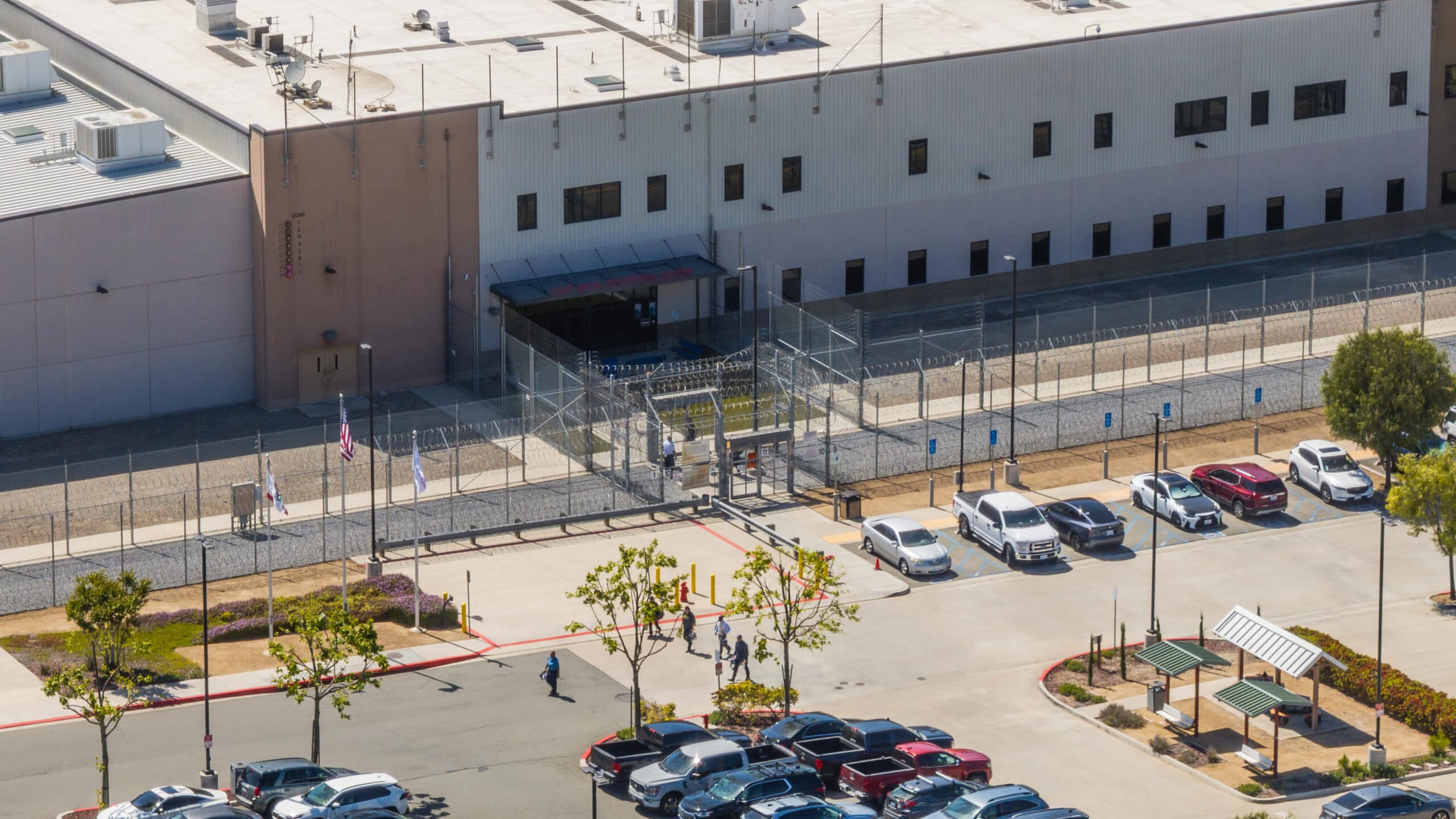 Aerial view of a fenced facility with armed security, several parked cars, people walking nearby, and a parking lot with trees and covered benches in the foreground.