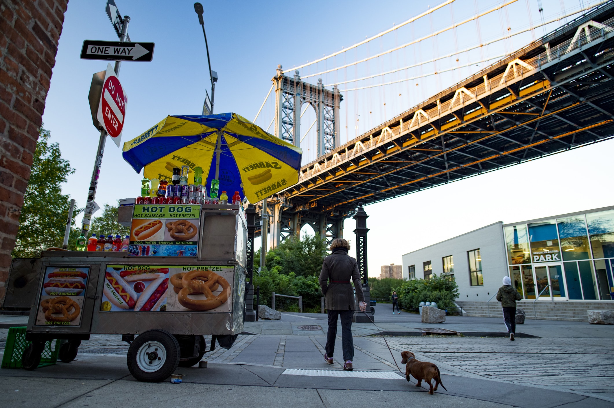 A person walks a small dog past a hot dog stand near a large suspension bridge in an urban setting. The stand displays snacks and drinks under a yellow and blue umbrella. Another person is in the background.