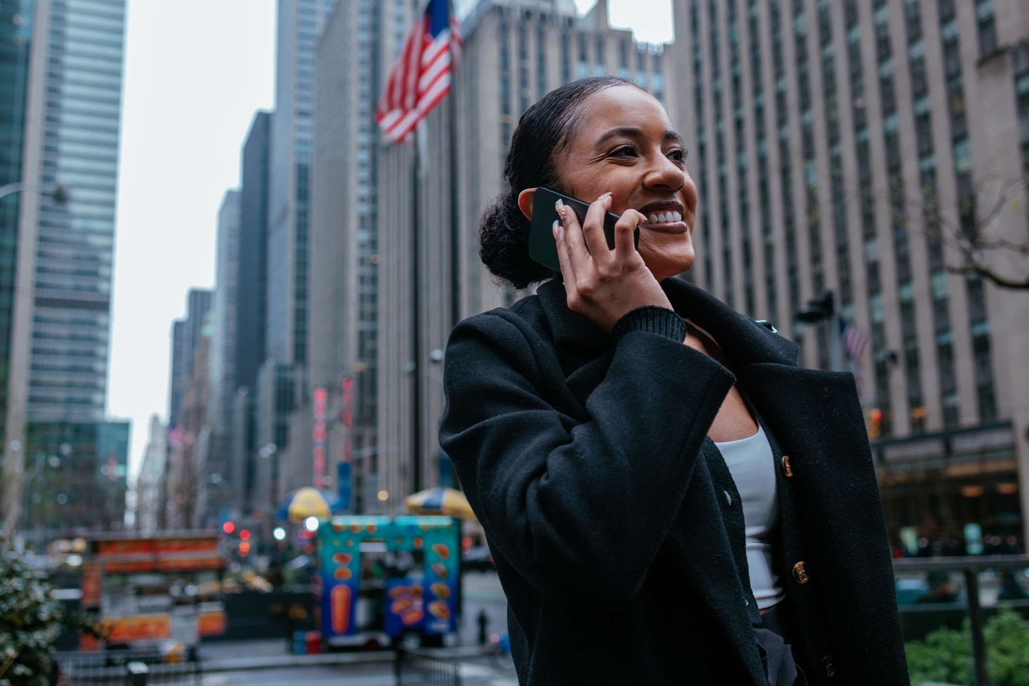 A woman in a dark coat smiles while talking on a smartphone outside in a city with tall buildings, an American flag, and food carts visible in the background.