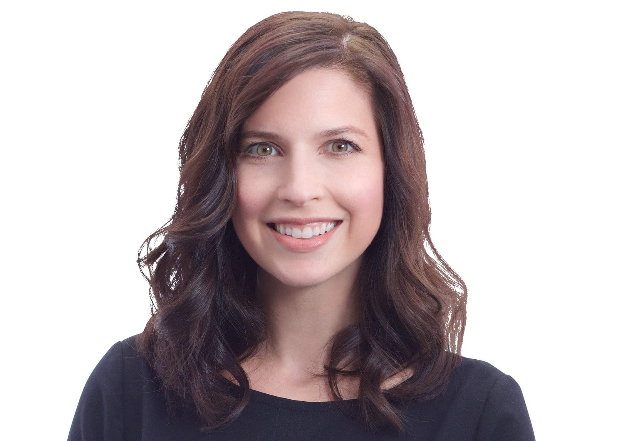 A woman with wavy brown hair, wearing a black top, smiles at the camera against a plain white background.