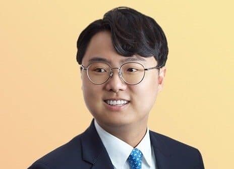 A man with short black hair and glasses is wearing a dark suit, white shirt, and blue tie with small patterns. He is looking slightly to the side and smiling. The background is a gradient of light yellow and peach.