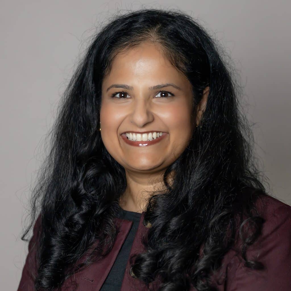 Headshot of a smiling woman with long, dark wavy hair wearing a burgundy jacket.