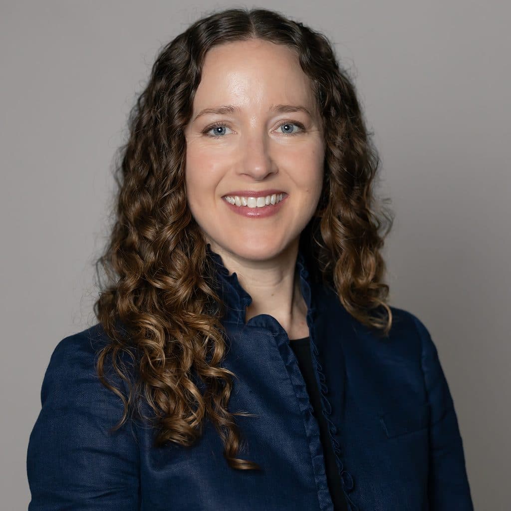 A woman with long, curly brown hair wearing a dark blue jacket smiles at the camera.