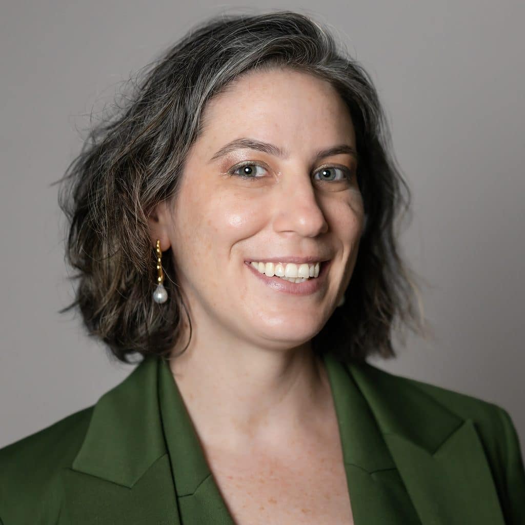 A smiling woman with shoulder-length grey hair wearing a green blazer and pearl drop earrings.