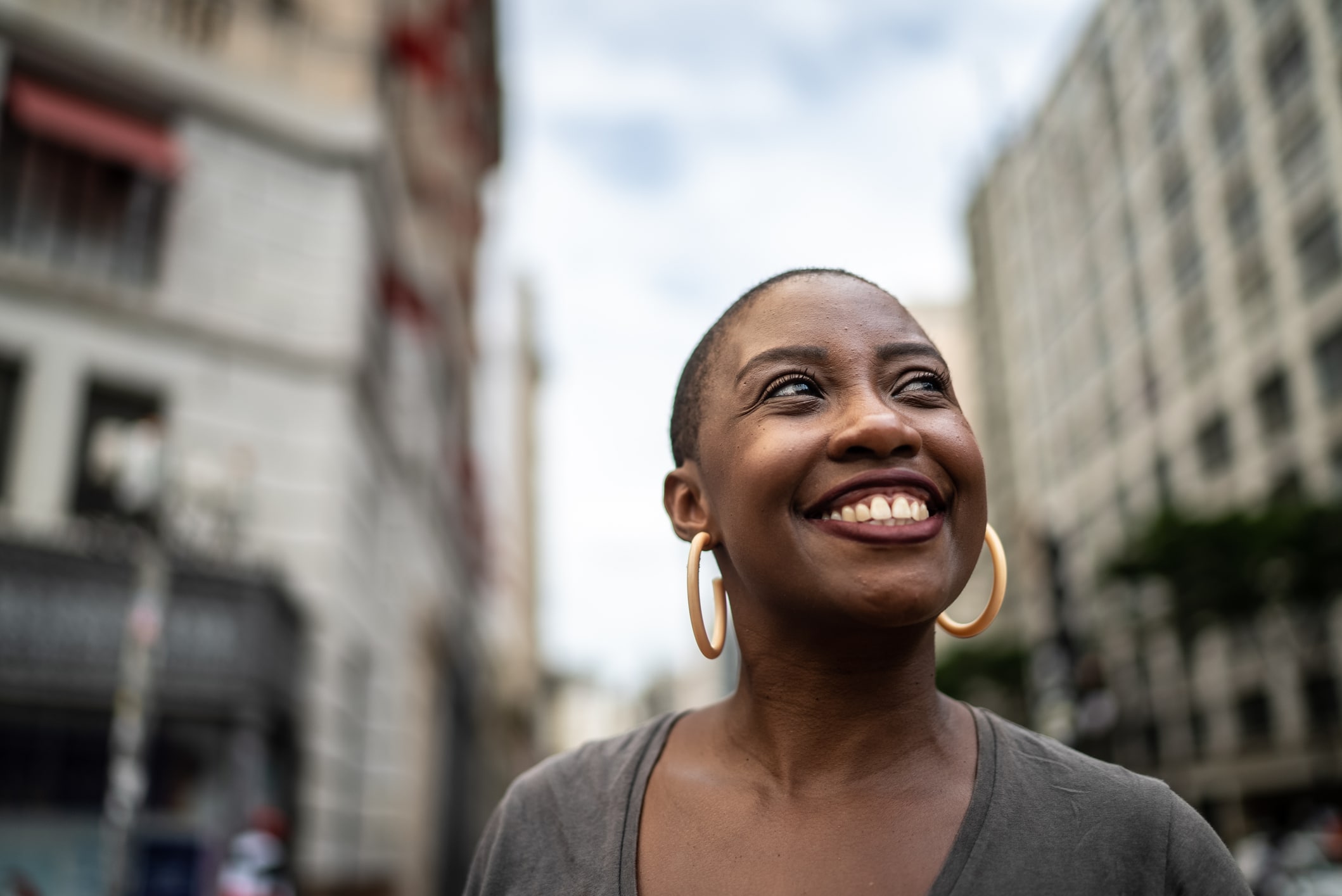 A person with short hair and large hoop earrings smiles while standing outdoors in an urban setting with tall buildings and a cloudy sky in the background.