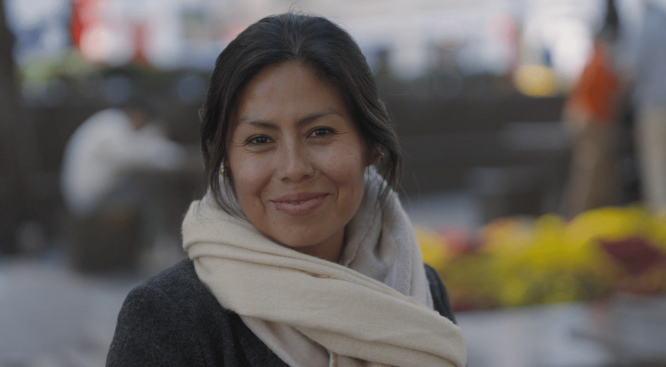 A woman with dark hair smiles at the camera, wearing a light scarf and dark coat. The background is blurred with people and colorful flowers, suggesting an outdoor location.