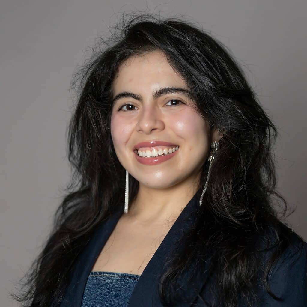 A young woman with long black hair smiles at the camera, wearing a navy blue blazer and long earrings.