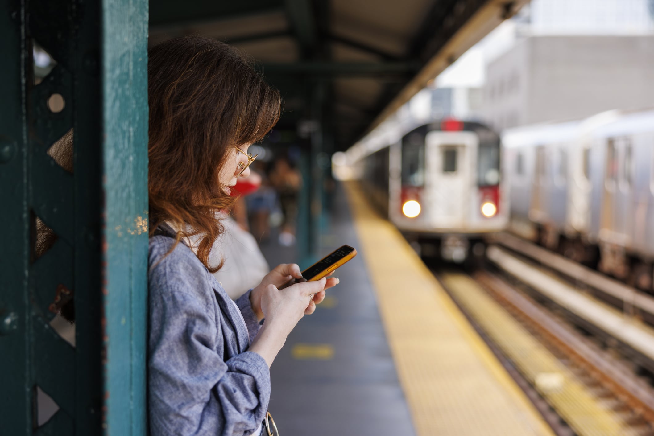 A woman in glasses and a blue shirt stands on a train platform, leaning against a support beam while looking at her phone. A subway train approaches in the background on a parallel track.
