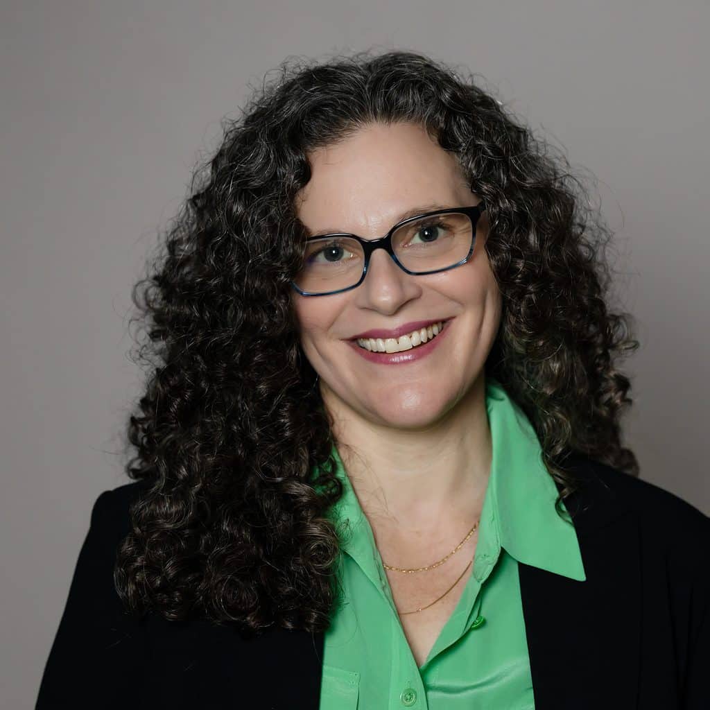 A woman with curly hair and glasses smiles while wearing a bright green button-down shirt and a black blazer.