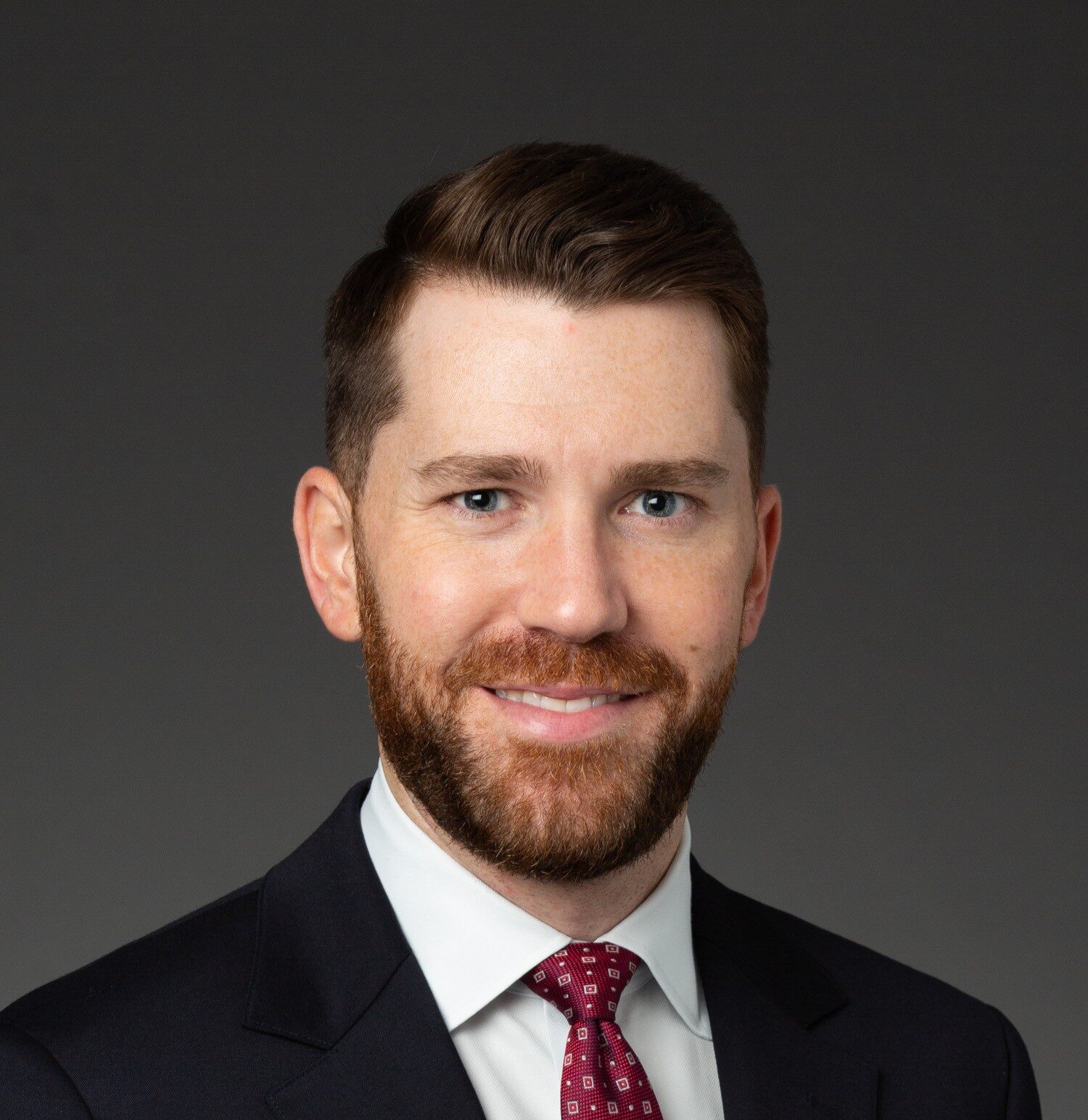 A man in a dark suit, white shirt, and red patterned tie poses against a plain dark background. He has short brown hair, a trimmed beard, and is smiling slightly while looking at the camera.