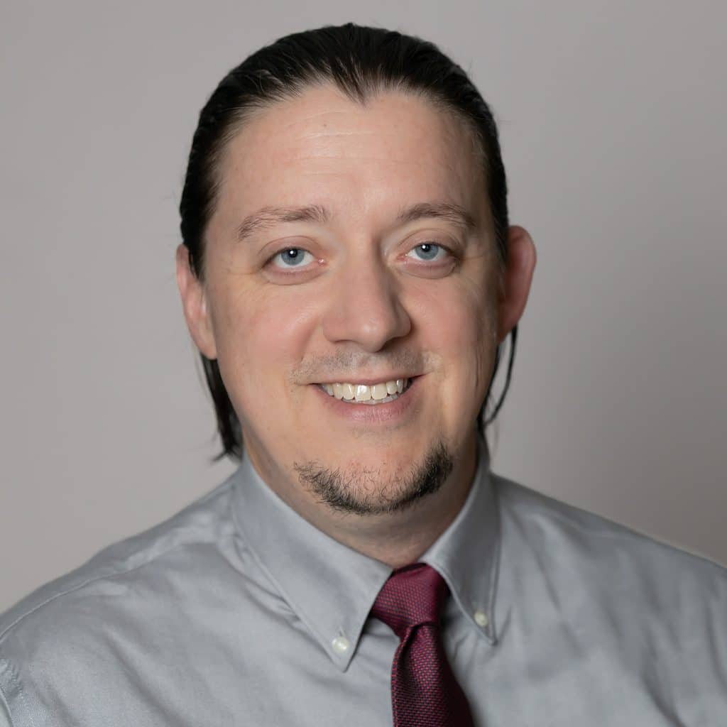 Headshot of a man with pulled-back hair and a goatee, wearing a light gray collared shirt and a dark red tie.