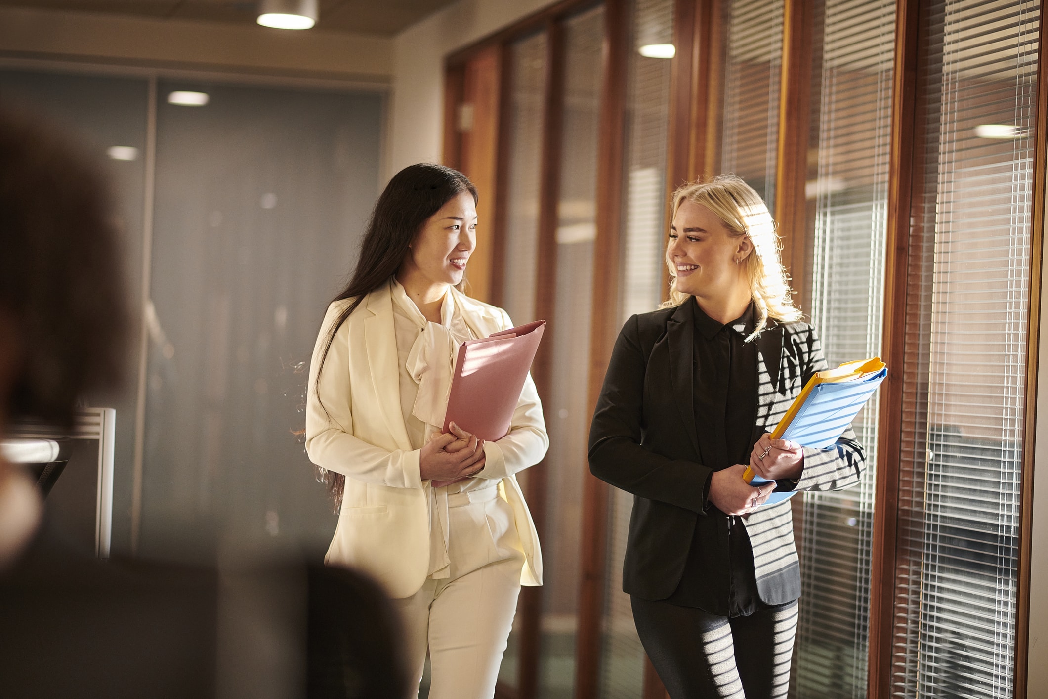 Two women in business attire walk and talk in an office hallway, holding folders and documents, with sunlight coming through window blinds beside them.