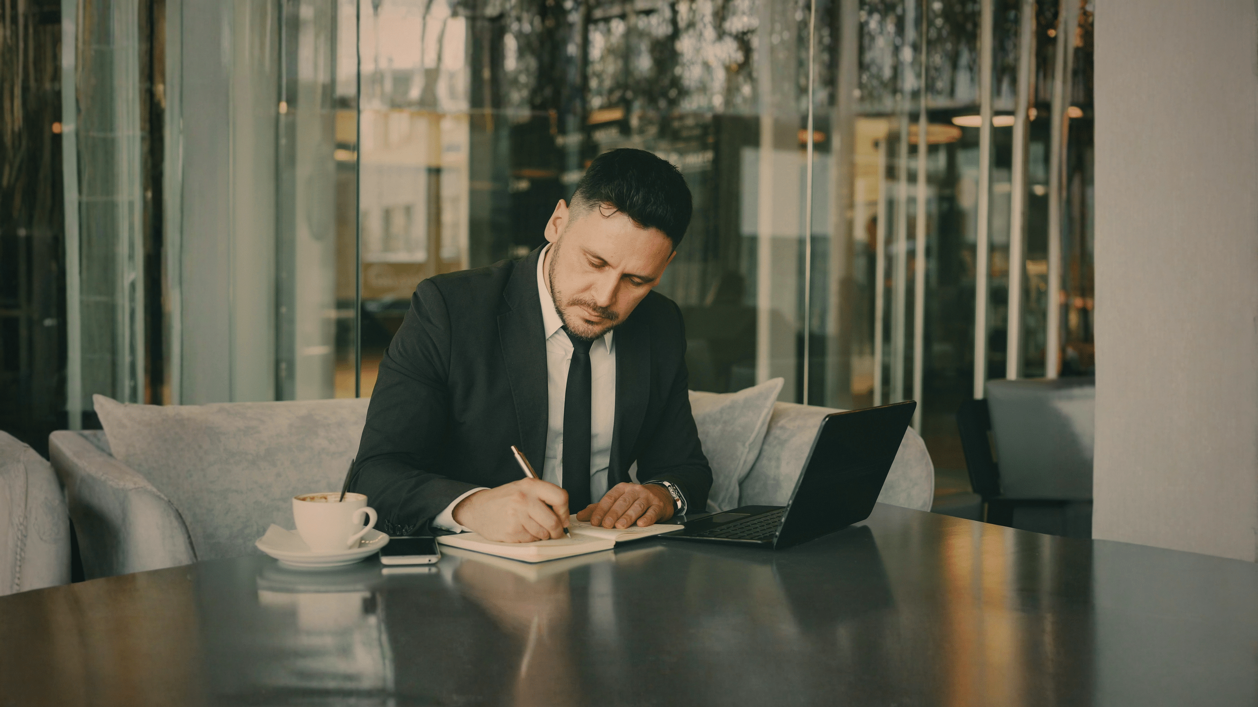 A man in a suit sits at a table with a laptop, notebook, and cup of coffee. He is writing in the notebook, with a phone placed nearby. The setting appears to be a modern office or café with large glass windows.