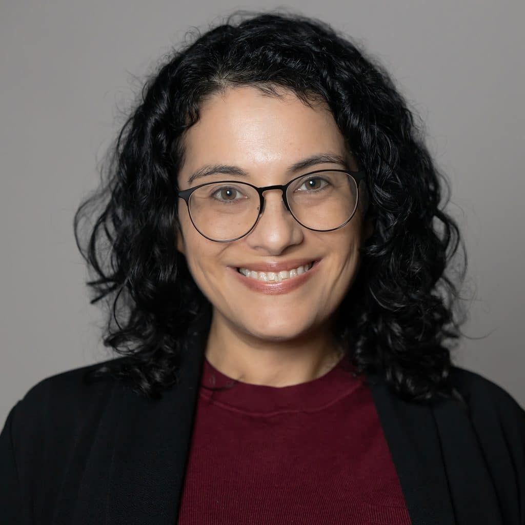 A woman with dark curly hair and glasses smiles in a professional headshot.
