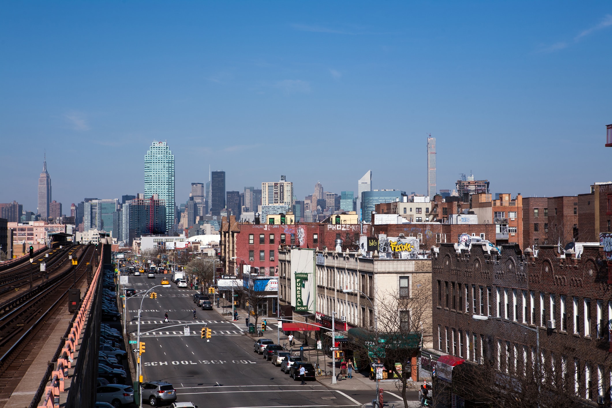 A cityscape shows a busy urban street with cars, brick buildings, shops, and elevated train tracks on the left. In the distance, skyscrapers are visible under a clear blue sky.