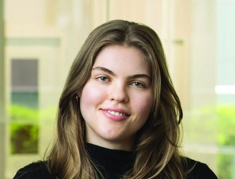 A young woman with long brown hair and a black short-sleeve shirt stands indoors in front of a window with greenery visible outside. She is facing the camera and smiling softly.