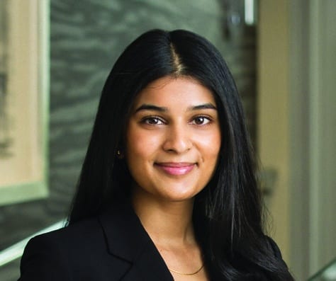 A woman with long dark hair, wearing a black blazer and a black top, stands indoors against a blurred background with neutral tones. She is facing the camera and smiling softly.