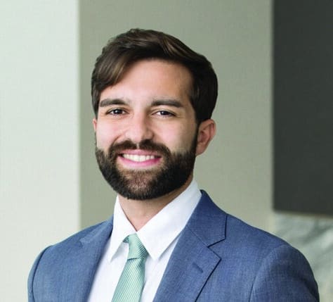 Man wearing a light blue suit, white shirt, and light green tie, standing indoors and smiling at the camera. The background is softly focused with neutral-colored walls and a small section of marble visible.