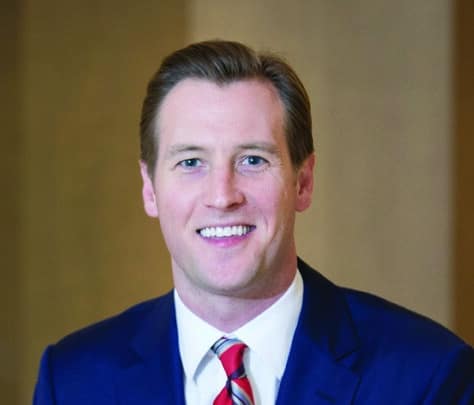 A man with short light brown hair wearing a blue suit, white shirt, and red striped tie is smiling and sitting in front of a blurred brown background.