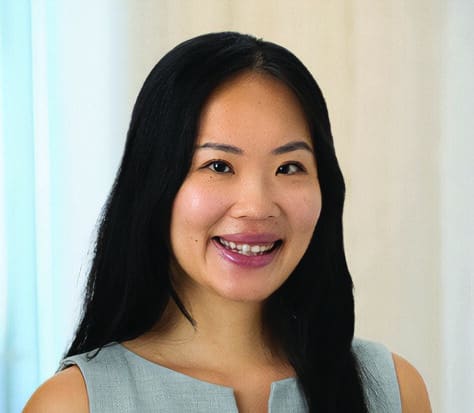 A woman with long black hair wearing a light gray sleeveless top stands in front of a softly blurred, light-colored background, smiling at the camera.
