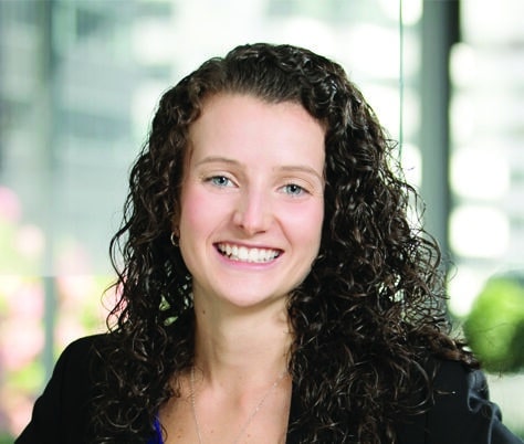 A woman with long curly hair wearing a blue blouse and black blazer smiles at the camera. She is standing indoors with large windows and a blurred background featuring greenery and buildings.