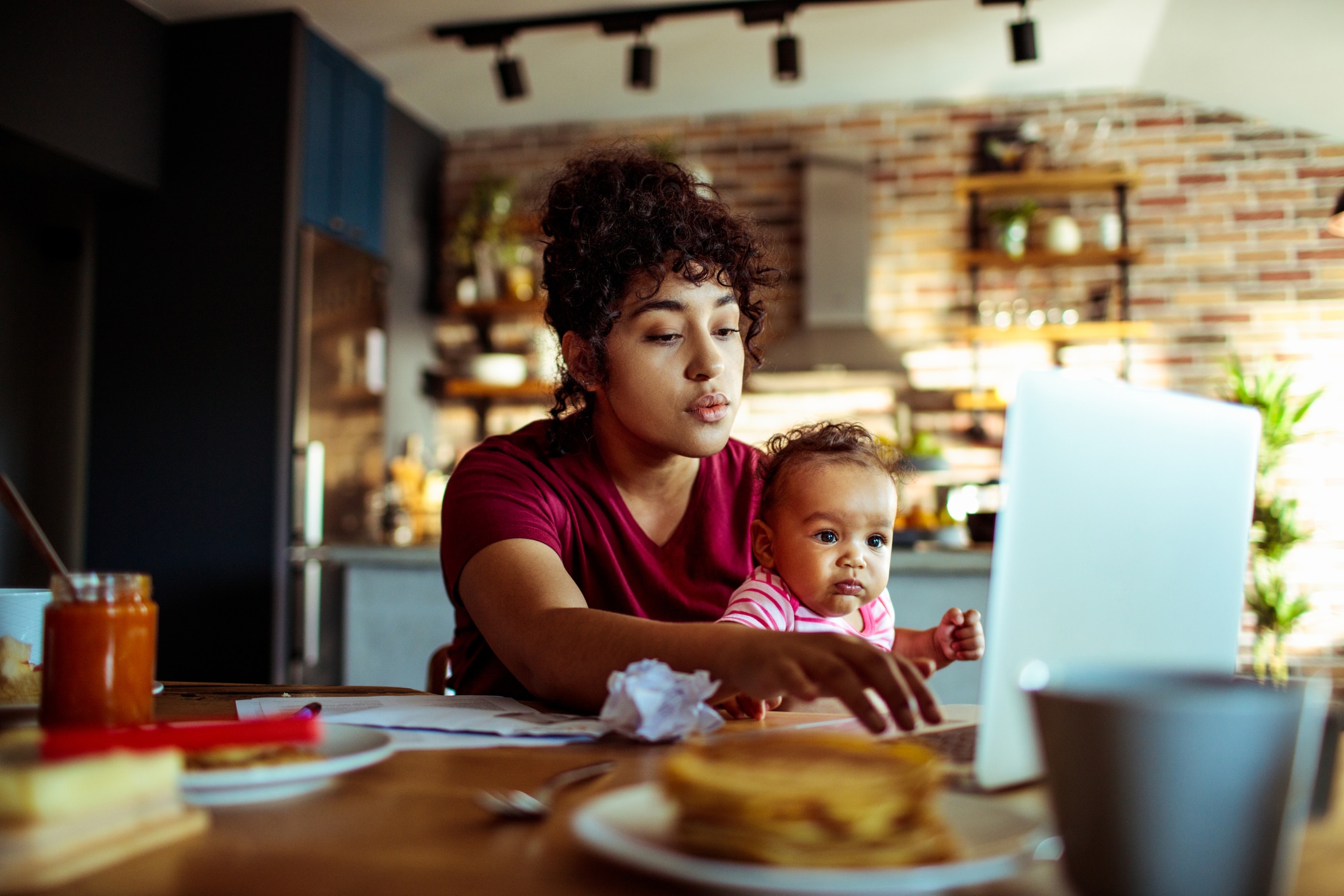A woman sits at a kitchen table using a laptop while holding a baby. Papers, food, and a coffee mug are on the table. The kitchen is visible in the background.