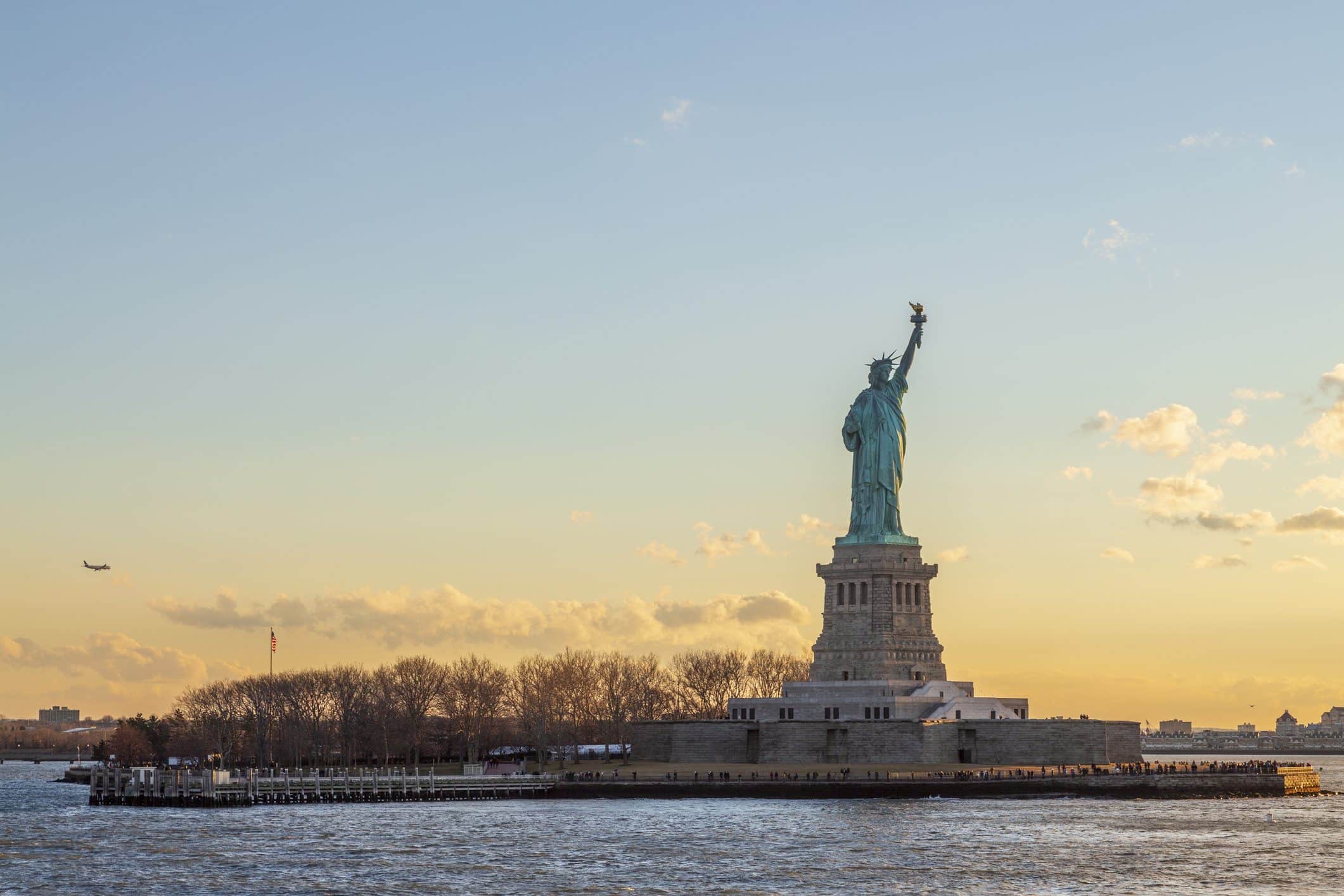 The Statue of Liberty stands on Liberty Island in New York Harbor at sunset, with trees and a pier in the foreground and a plane visible in the sky to the right.