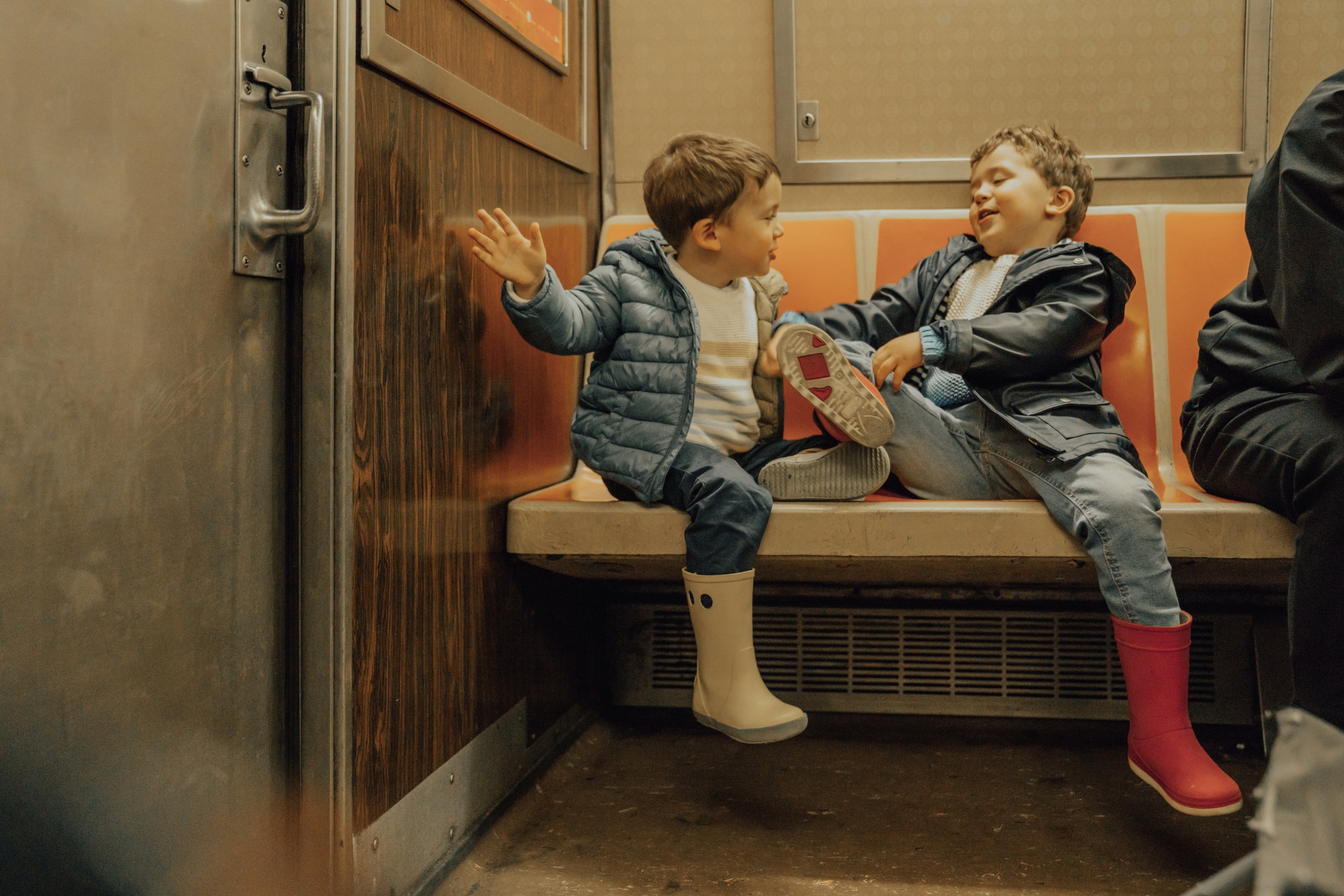 A woman sits on a subway bench, holding a small child in her lap. Both appear relaxed. The subway doors behind them have warning signs, and another person is partially visible to the right.