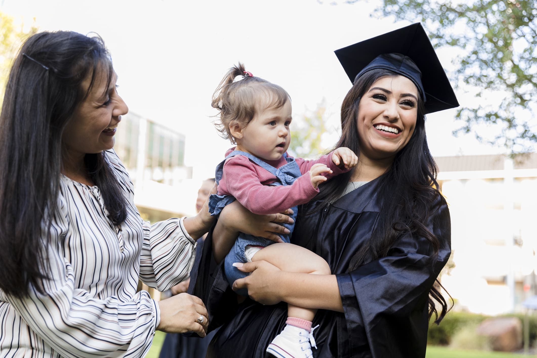 A woman in a graduation cap and gown holds a toddler while standing outside.