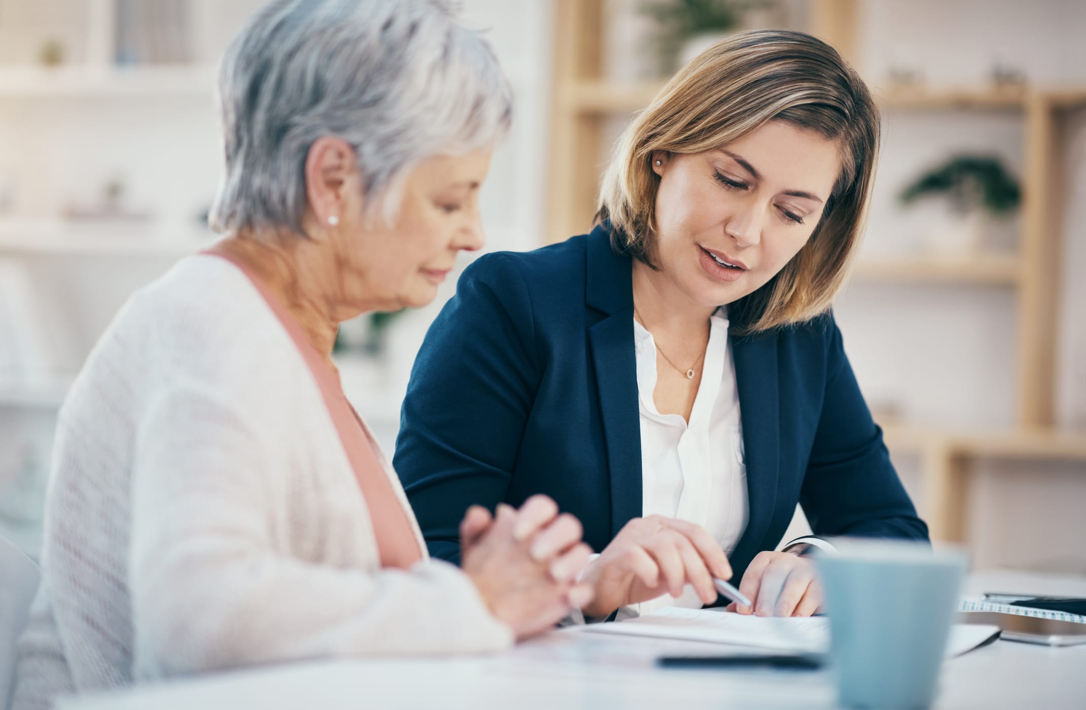 A younger woman in a suit explains paperwork to an older woman with gray hair. They are sitting together at a table in a bright room with shelves and plants in the background.
