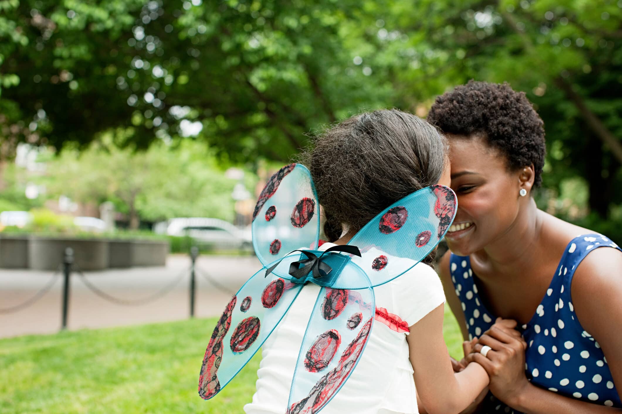 An adult and a child with ladybug wings touch foreheads and smile while kneeling on grass in a park. Trees and blurred buildings are visible in the background.